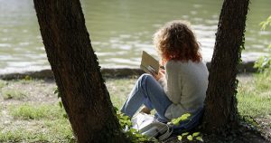 woman leaning against a tree next to a water front, reading a book
