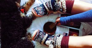 Girl in fuzzy slippers holding a book and a mug of hot chocolate with a small dog sleeping next to her
